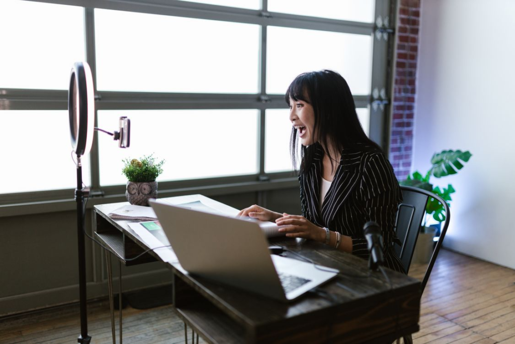 A woman sitting at a desk in front of a laptop and ring light setup, reacting expressively while recording content.